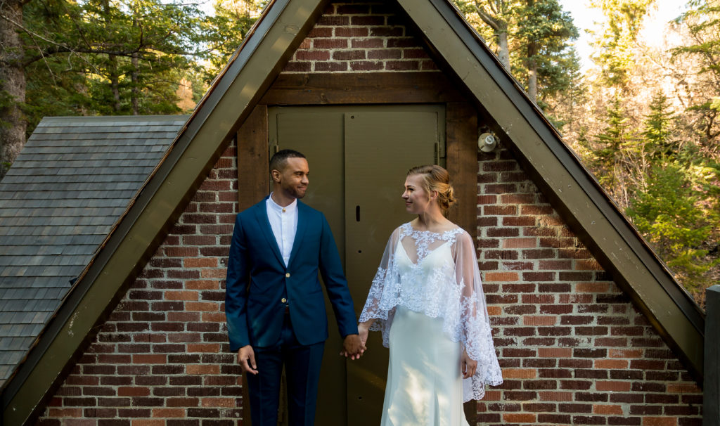 Bride and groom looking at each other while holding one hand in the middle of them with their bodies facing the camera and standing in front of a cabin in the woods.