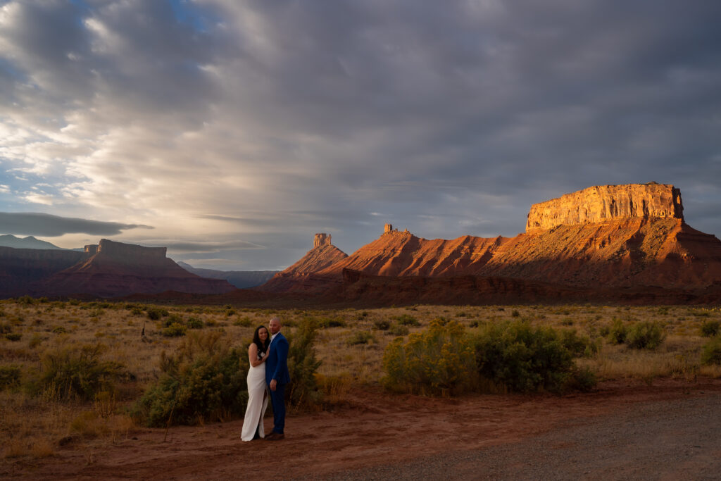 Bride and groom with bodies facing each other and looking at the camera. Bride has one hand on groom's chest and the other wrapped around his waist. Groom has back hand around bride's waist and the other hand hanging down in front. Couple is standing in front of large buttes that have the sun hitting them as it rises creating an alpenglow in Moab, Utah