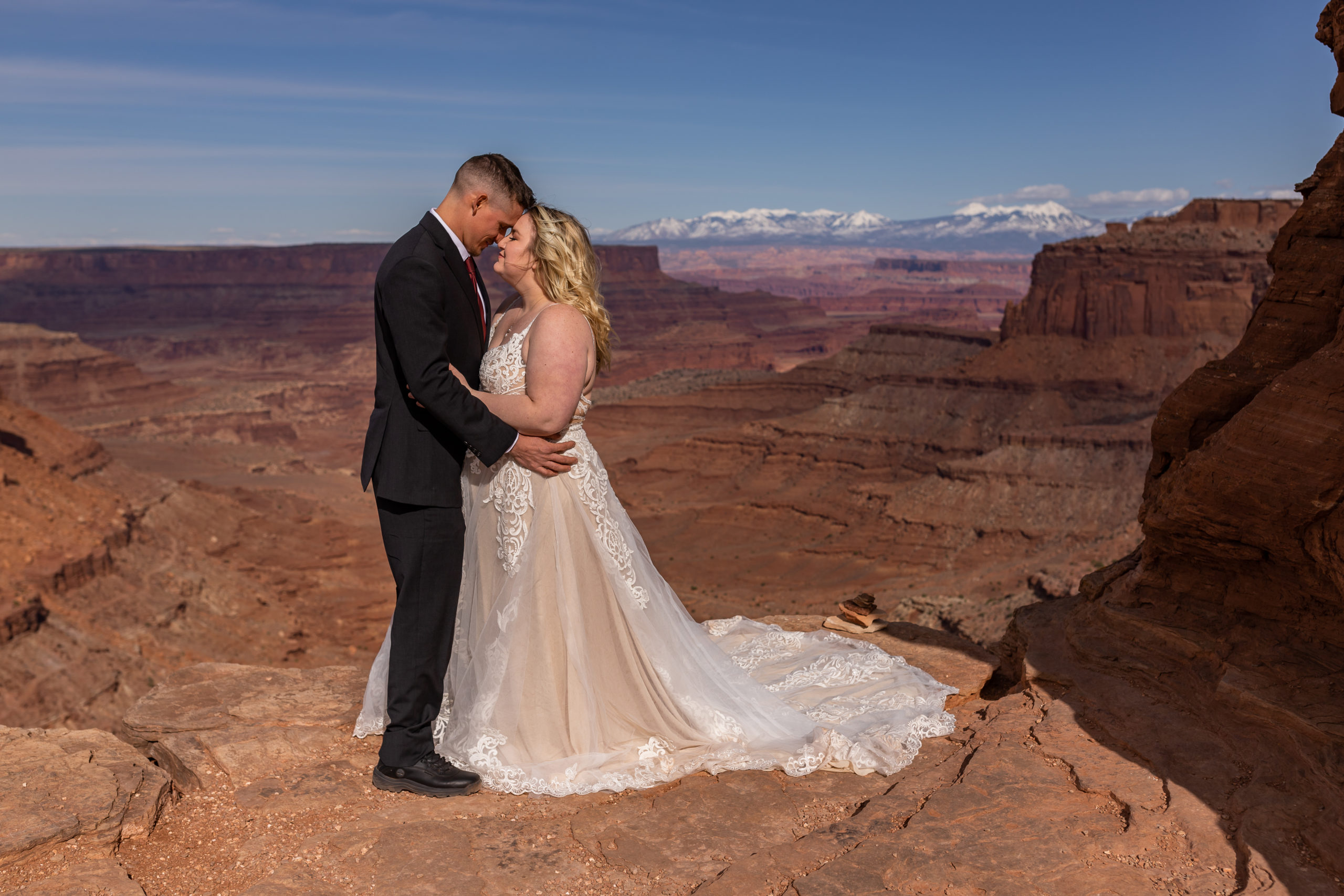Bride and groom stand on cliff edge overlooking red sandstone canyon below in Canyonlands National Park. It is a clear day with blue skies and you can see the La Sal mountains way in the distance.