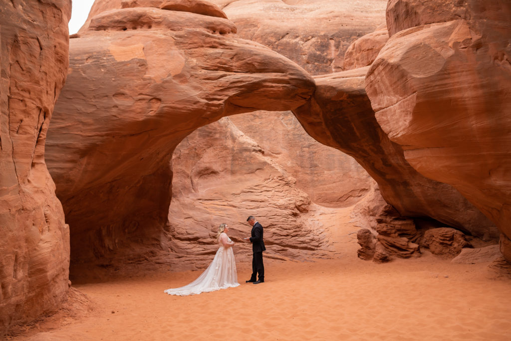 Bride and groom standing facing each in front of Sand Dune Arch in Arches National Park in Moab, Utah