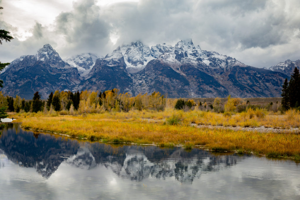 The Teton Mountain Range with gloomy clouds over the peaks in the fall with the mountains reflection in the water in front of them