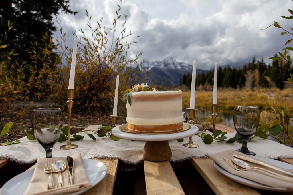 A close up photo of the cake from the side. It has off white frosting and is a naked cake with some of the actual cake showing through the frosting. It also has real flowers that are red, yellow, orange, white, and green on top of it. You can see the Teton mountain range in the back and the cake is shown with the table set up
