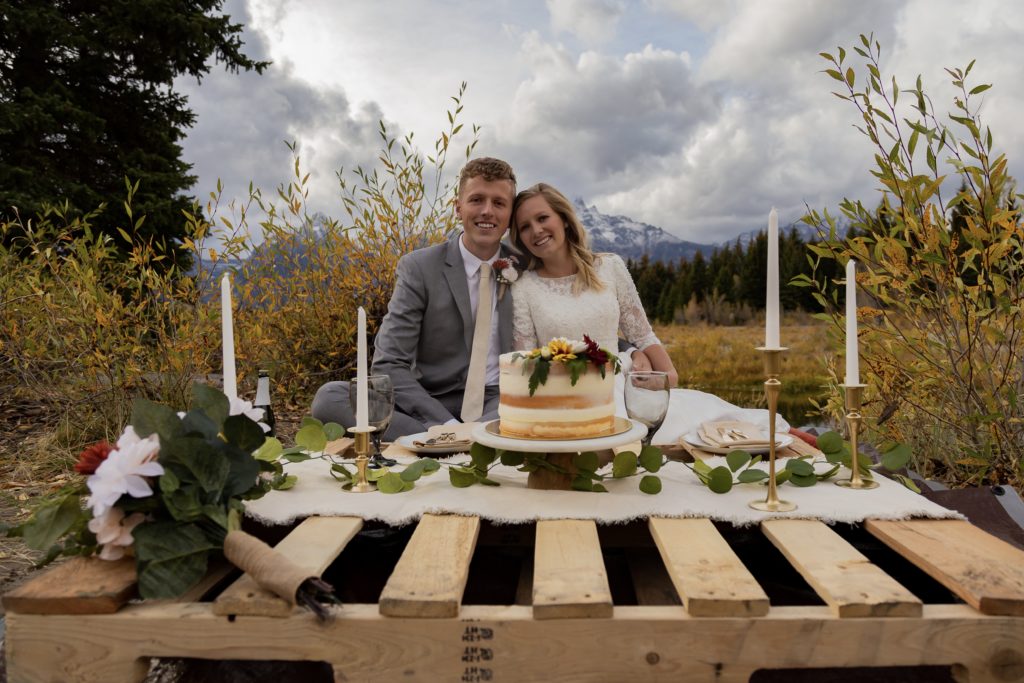 Grand Teton National Park Elopement. Bride and groom sitting down at a picnic with cake and sparking cider. There are candles and flowers around the cake. The Teton mountain range is peaking out behind them It is fall time and there are yellow and green trees and bushes.