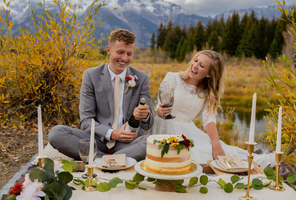 Grand Teton National Park Elopement. Bride and groom sitting down next to each other at a picnic with cake and sparking cider. They are laughing as groom opens the cider. There are candles and flowers around the cake. The Teton mountain range is peaking out behind them It is fall time and there are yellow and green trees and bushes.
