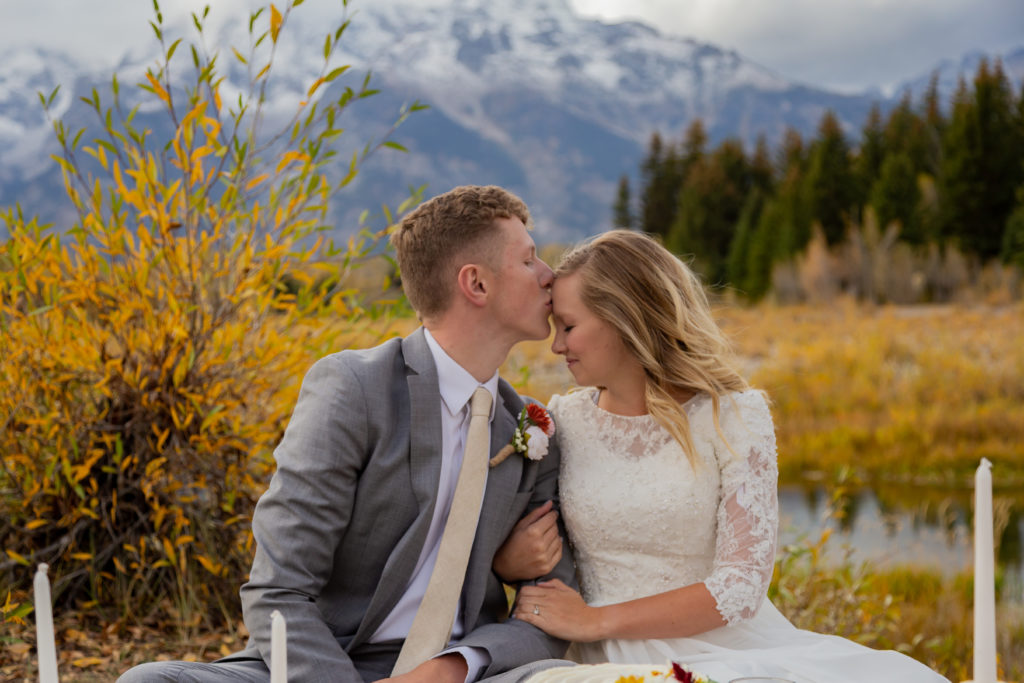 Grand Teton National Park Elopement. Bride and groom sitting down at a picnic. Groom is kissing bride's forehead. Bride has eyes closed taking in the moment. The Teton mountain range is peaking out behind them It is fall time and there are yellow and green trees and bushes.