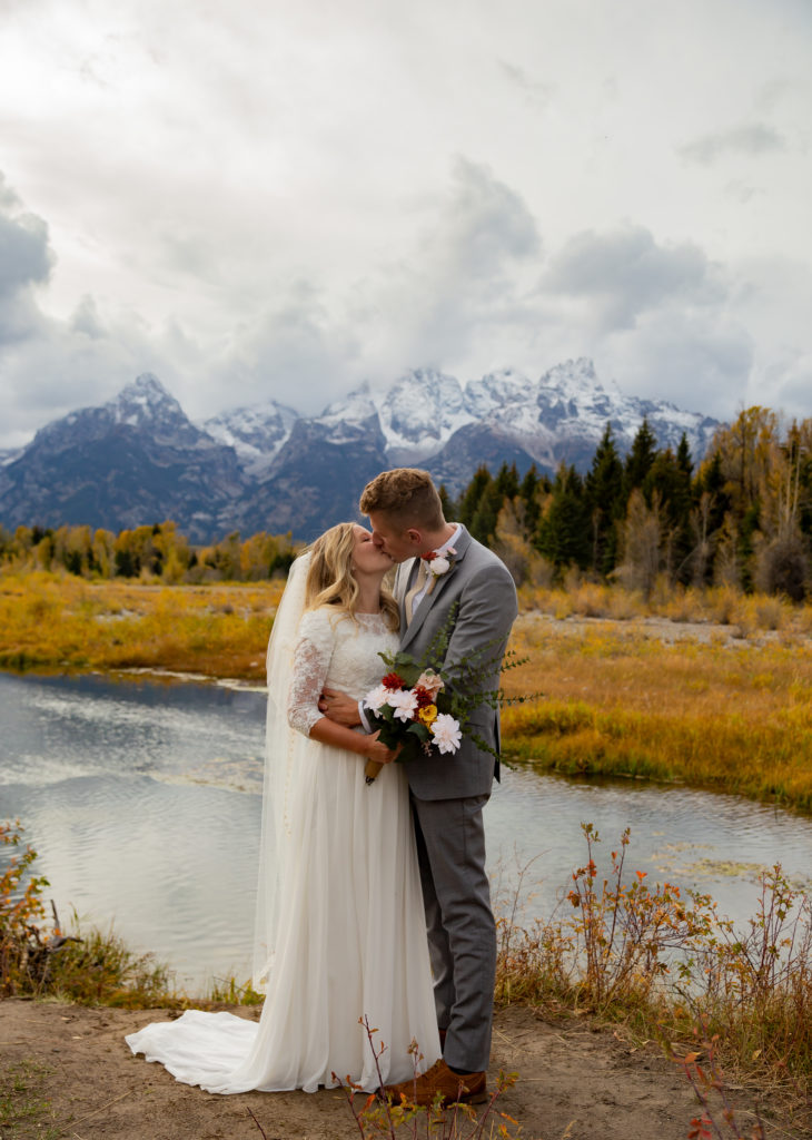 Grand Teton National Park Elopement. Bride and groom facing each other and kissing with the Teton mountain range behind them reflecting in the water that is behind them as well. It is fall time and there are yellow and green trees.