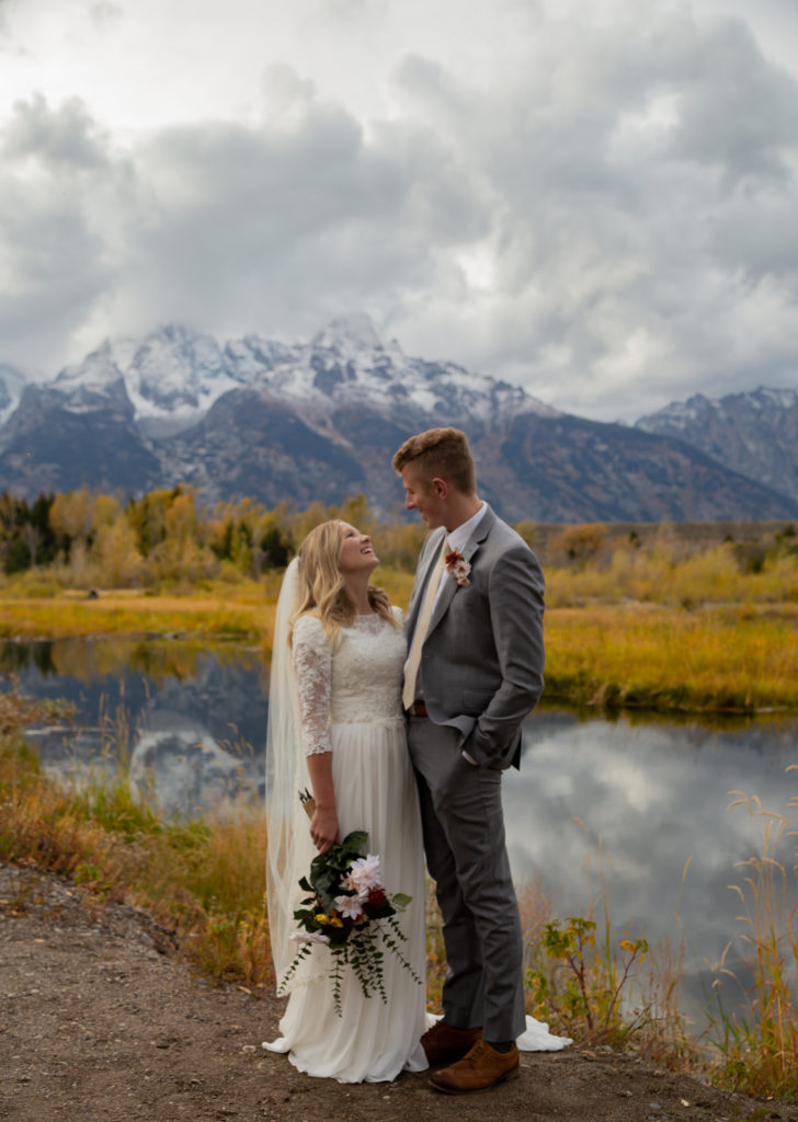 Grand Teton National Park Elopement. Bride and groom facing and looking at each other with the Teton mountain range behind them reflecting in the water that is behind them as well. It is fall time and there are yellow and green trees.