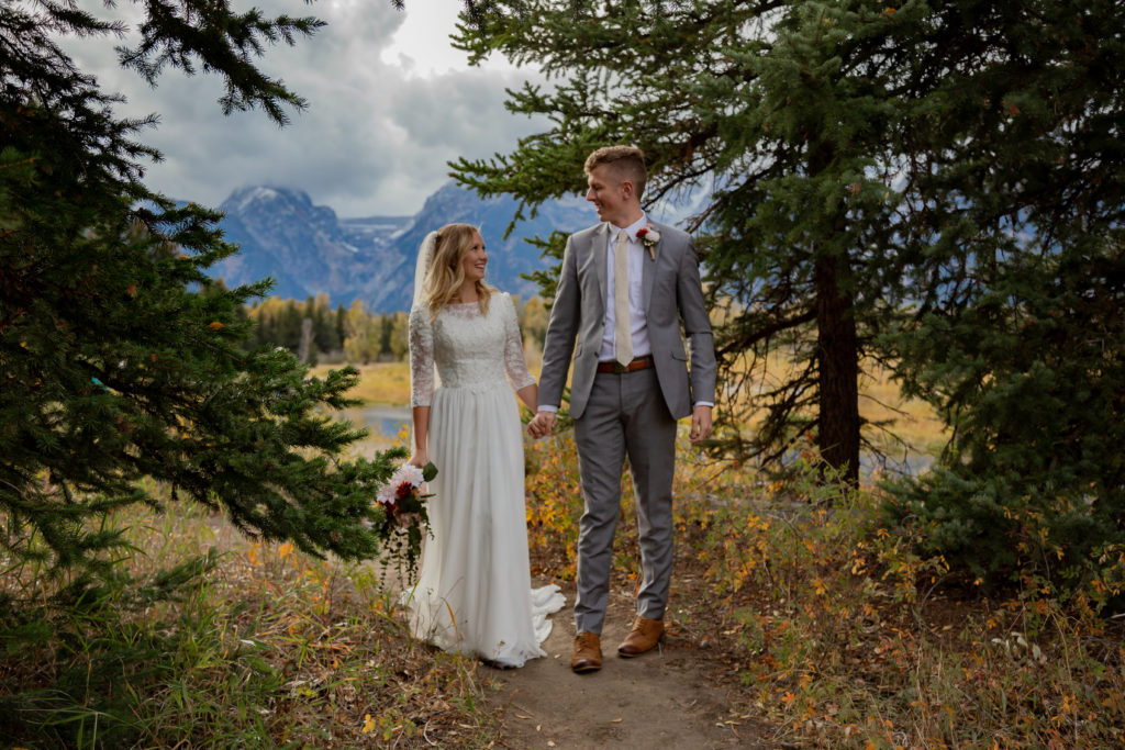 Bride and groom walking on a trail through pine trees with the Teton mountain range peaking through in the background in Grand Teton National Park in Wyoming.