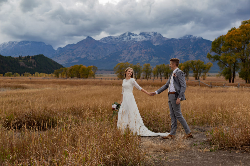 Grand Teton National Park Elopement. Bride leading groom through a field with the Teton mountain range behind them in the distance, looking back at him and they are smiling at each other.