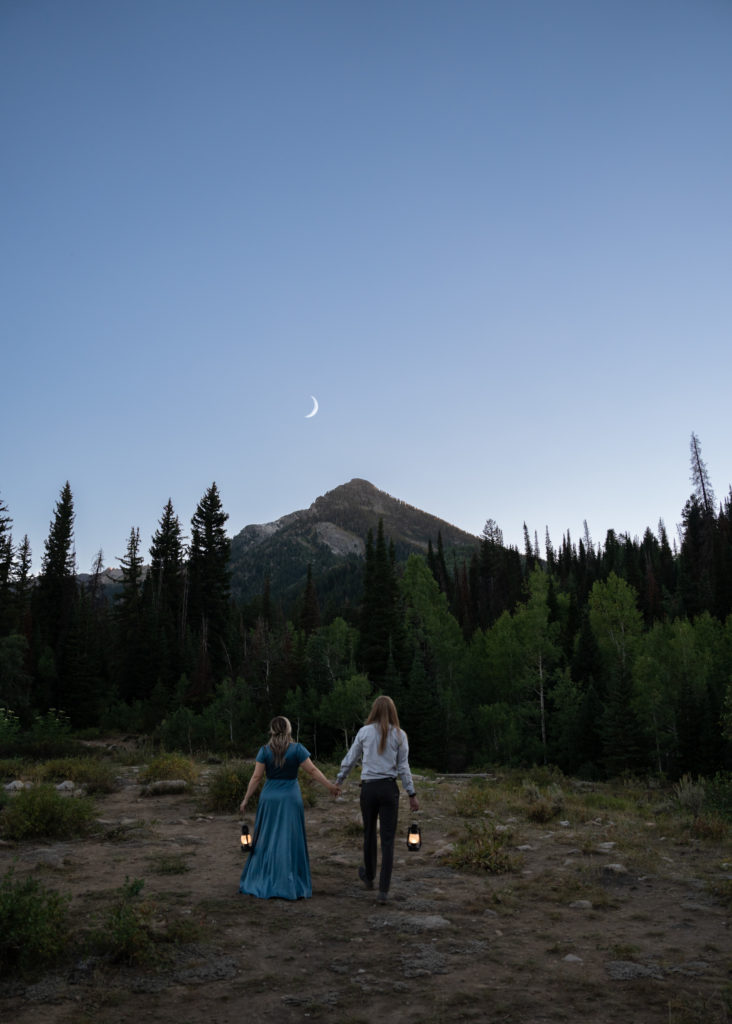 Couple walking away from camera towards trees holding lanterns at blue hour in Big Cottonwood Canyon in Salt Lake City, Utah