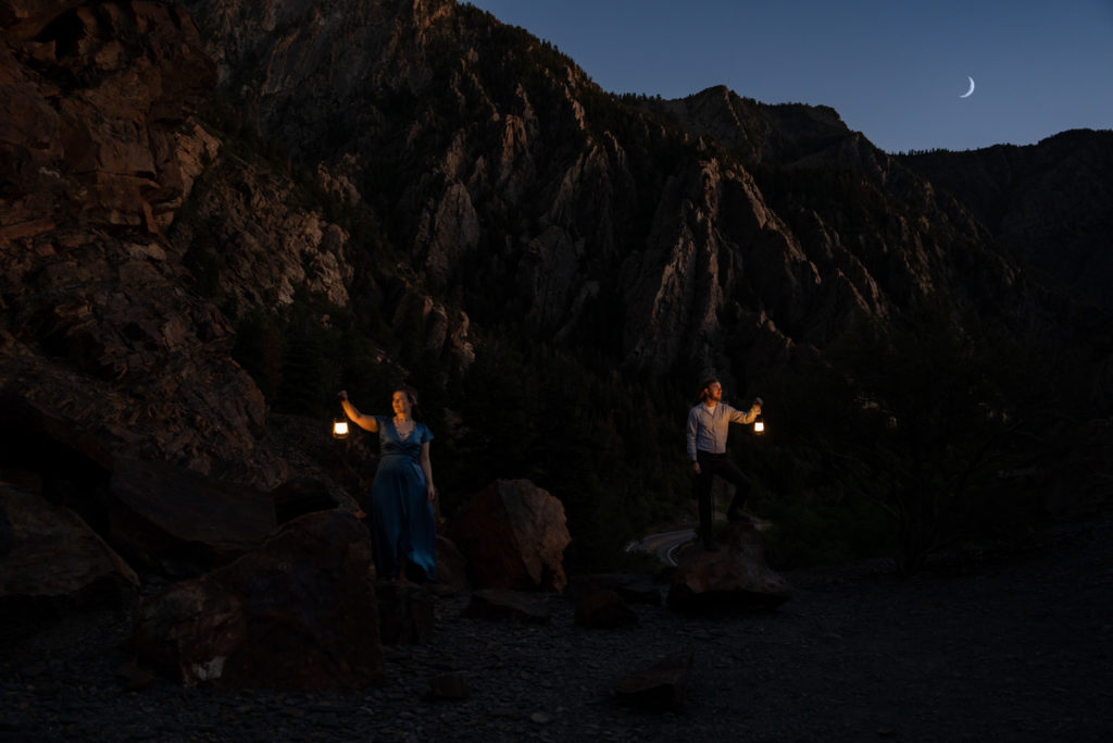 Couple standing on top of rocky mountain looking away from each other holding lanterns under the moon in Big Cottonwood Canyon in Salt Lake City, Utah