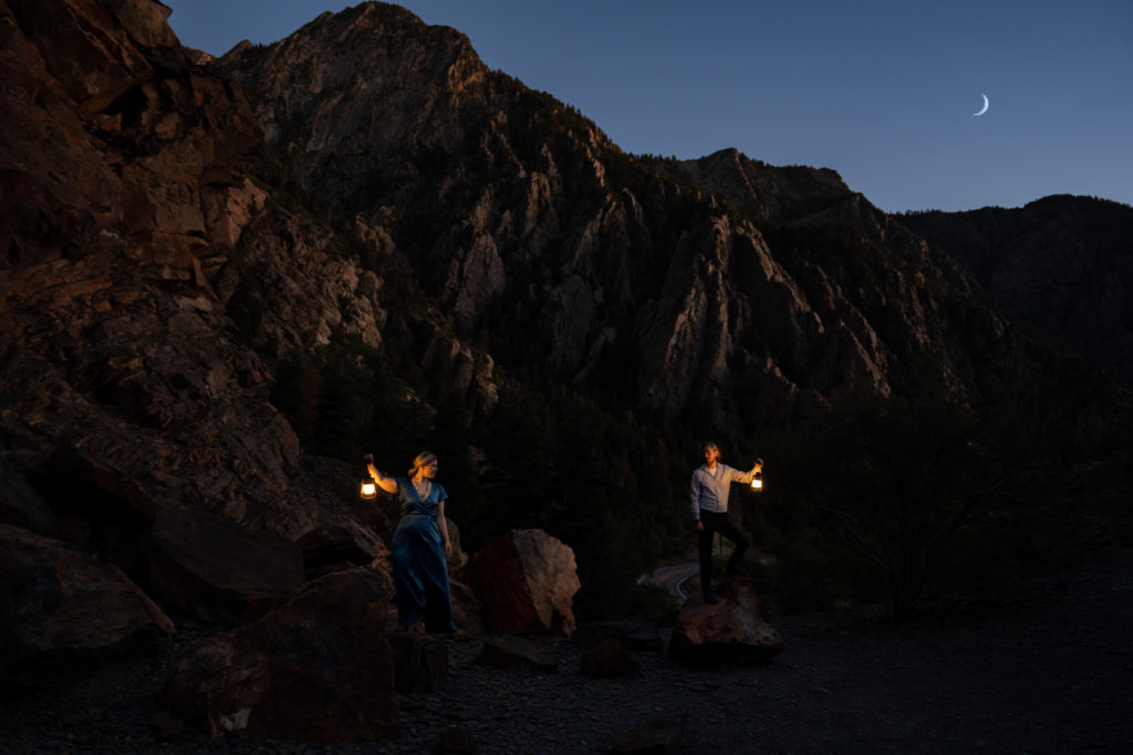 Couple standing on top of rocky mountain looking towards each other holding lanterns under the moon in Big Cottonwood Canyon in Salt Lake City, Utah