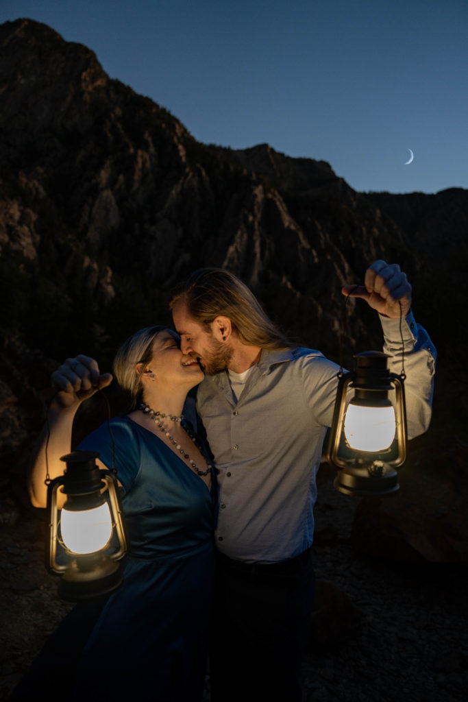 Couple standing on top of rocky mountain kissing holding lanterns in under the moon in Big Cottonwood Canyon in Salt Lake City, Utah