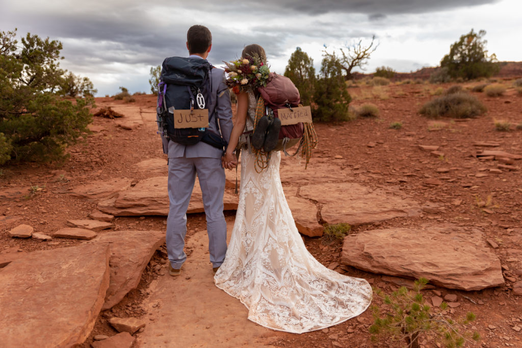 Bride and groom facing away from camera in wedding attire with their climbing gear on their backs with "just married" signs on their backpacks in Capitol Reef National Park