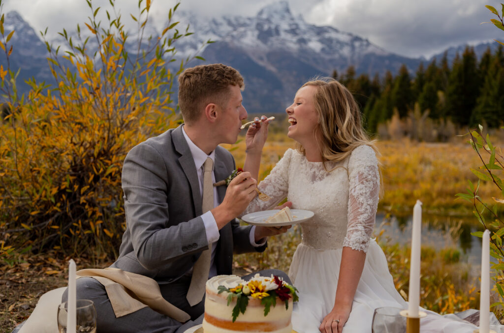 Fall Elopement in Grand Teton National Park in Jackson Hole, Wyoming. Bride is feeding her groom a piece of wedding cake and the couple is laughing together with the Teton mountains in the background.