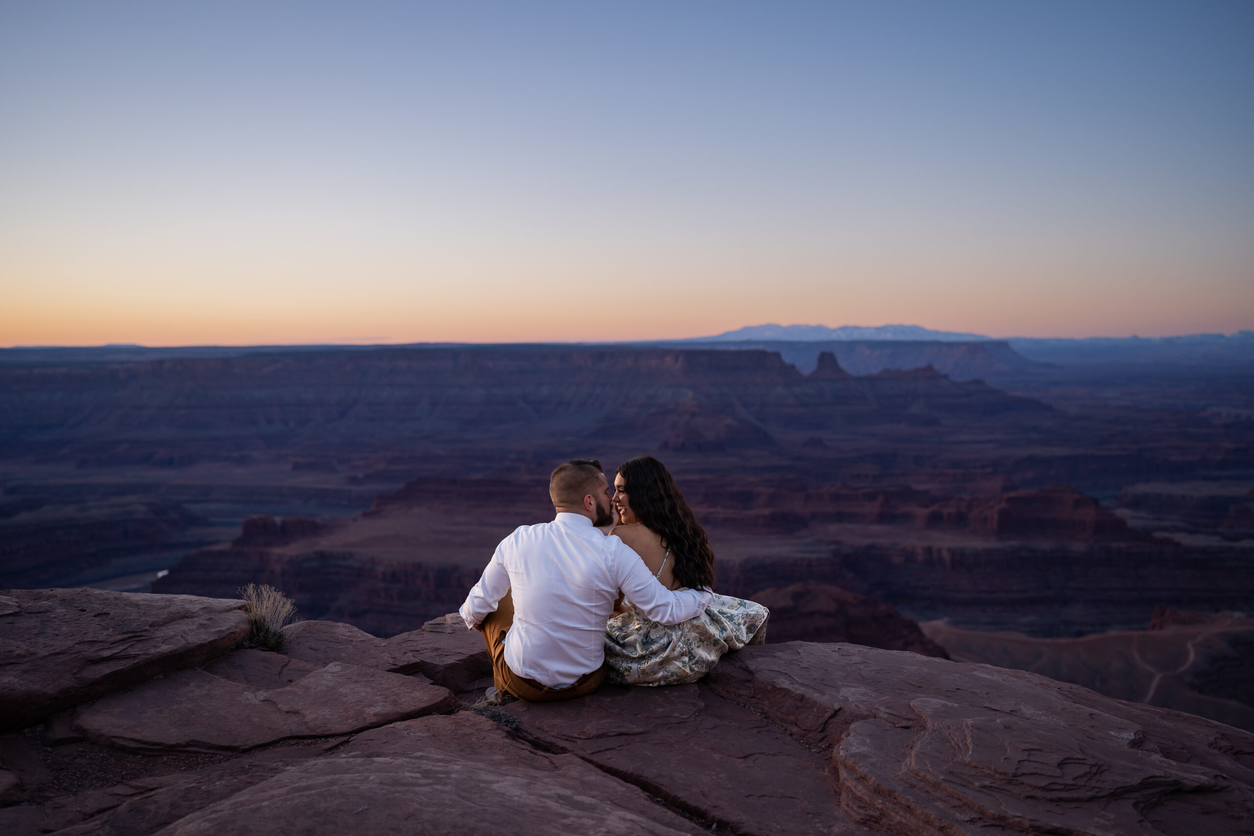 Sunrise elopement at Dead Horse Point in Moab, Utah with bride and groom sitting on the edge of a red rock cliff and kissing.
