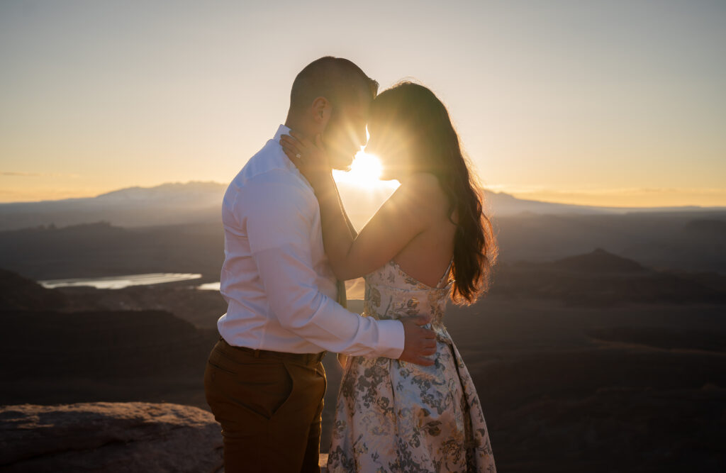 Bride and groom holding each other with their foreheads touching and the sun coming through between them during sunrise elopement at Dead Horse Point in Moab, Utah holding each other while the sun comes through between them as it rises over the mountains behind them.