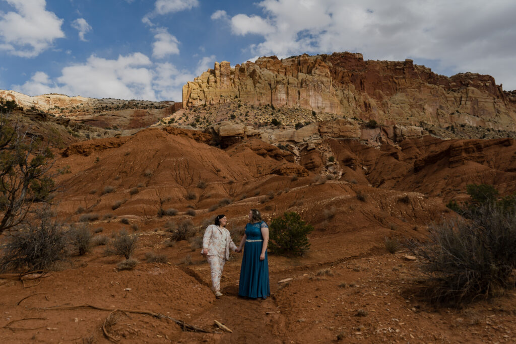 Two brides walking hand in hand down a trail with red rock walls all around them in Capitol Reef National Park in Utah.