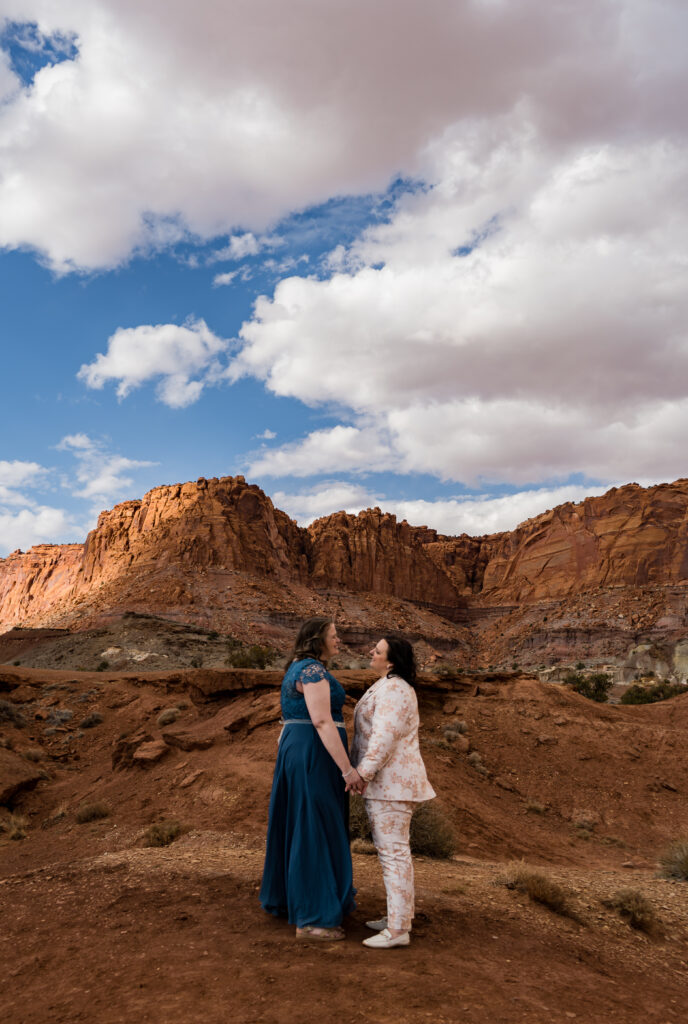 Two brides facing each other and holding hands between them with red rock walls and towers behind them in Capitol Reef National Park in Utah.