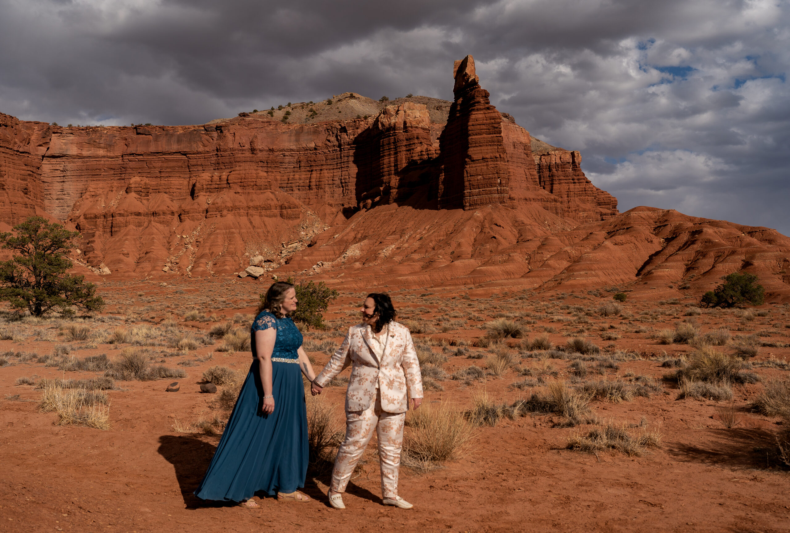 One bride is leading the other bride across the photo while holding hands with red rock walls and towers behind them in Capitol Reef National Park in Utah.