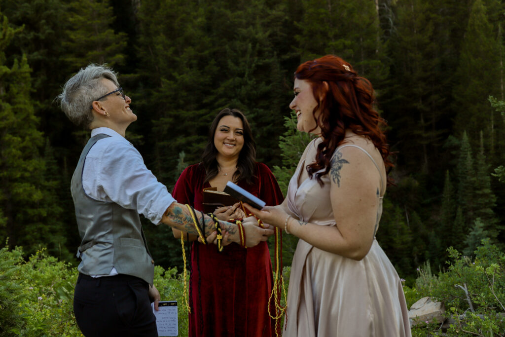LGBTQIA+ mountain elopement in Salt Lake City, Utah with two brides standing in front of their officiant during their hand fasting ceremony and everyone is laughing.