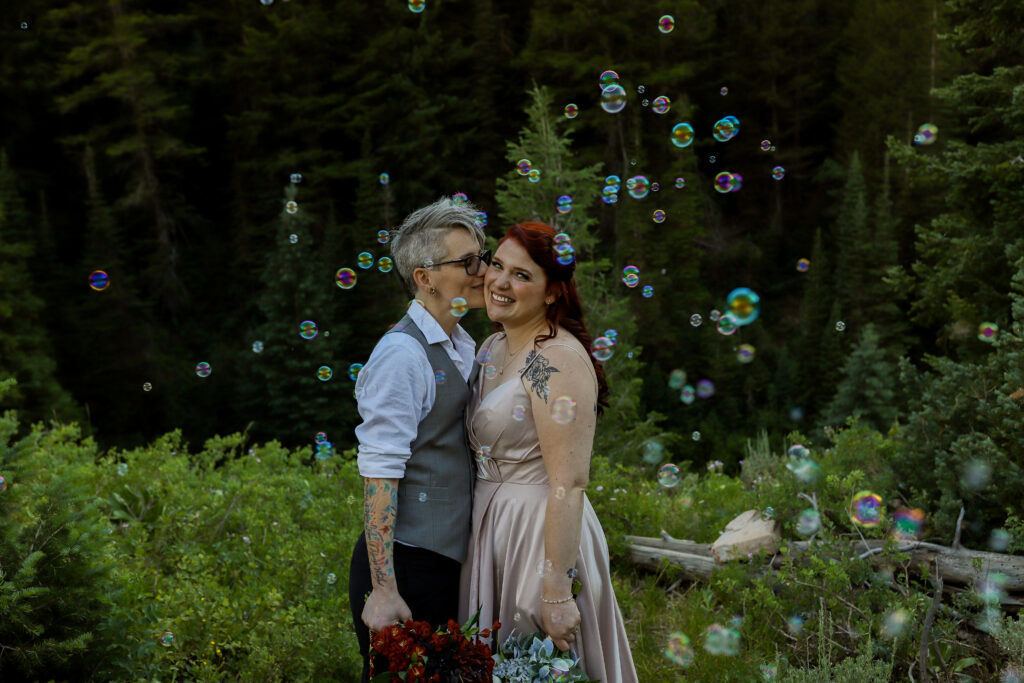 LGBTQIA+ mountain elopement in Salt Lake City, Utah with two brides side hugging and one kissing the other on the cheek while the other one is smiling at the camera with bubbles being blown in front of them.