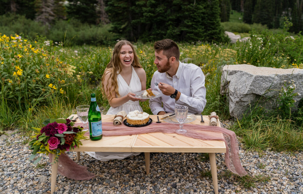 Bride and groom sitting down at a picnic table to celebrate being married with a charcuterie board and cake. Bride is laughing and groom is smiling at her.