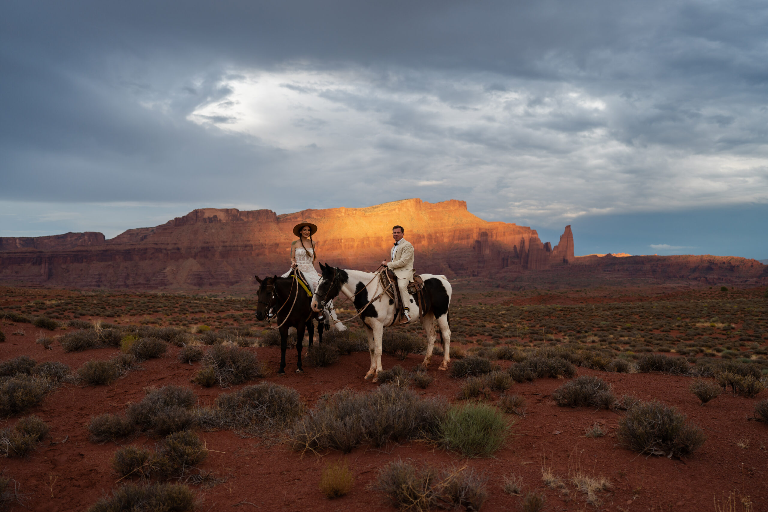 Bride and groom on horses facing the camera with the sun setting on the buttes and cliffs in the background in Moab, Utah