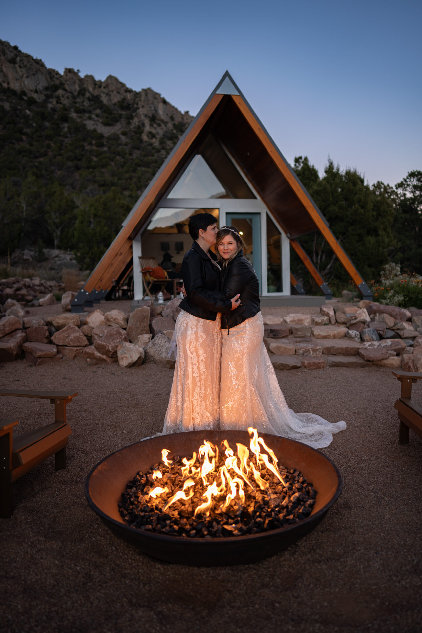 Two brides embracing each other in front of a fire wearing their dresses and matching black leather jackets in front of their A-Frame cabin in Colorado.