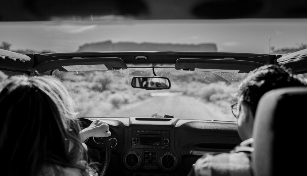 Black and white photo of two brides in the front seats of a Jeep. Photographer is sitting in the back taking photo from behind the couple.