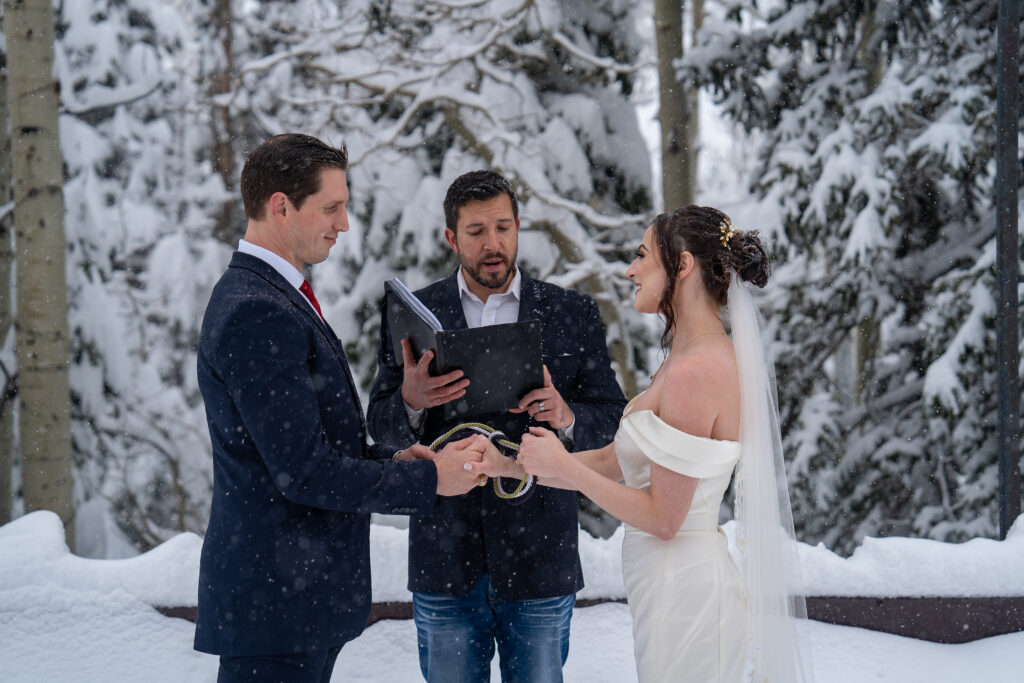 Bride and groom doing a hand fasting ceremony in front of their officiant while snow is falling and they are surrounded by snow covered trees.