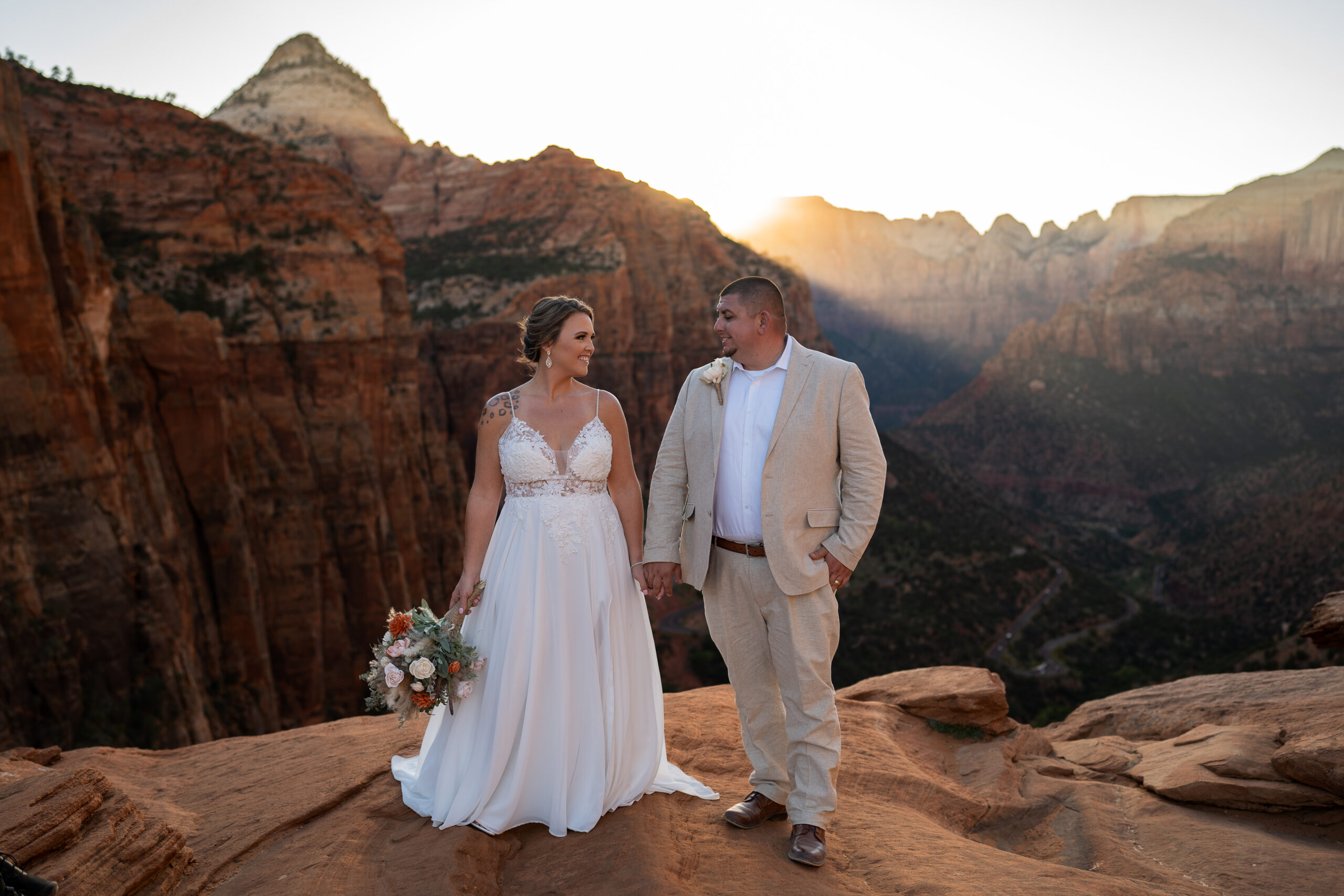 Elopement with Bride and Groom standing apart with their bodies facing the camera holding hand in the middle of the two of them looking at each other in Zion National Park in Utah at Canyon View Overlook at Sunset