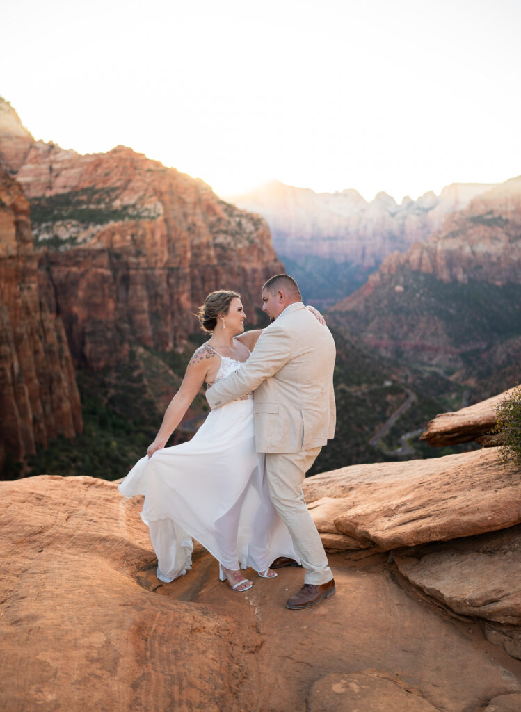 Elopement with Bride and Groom facing each other. Bride is holding out her dress with one hand and has the other wrapped around the groom's neck. Groom is holding bride around the waist in Zion National Park in Utah at Canyon View Overlook at Sunset