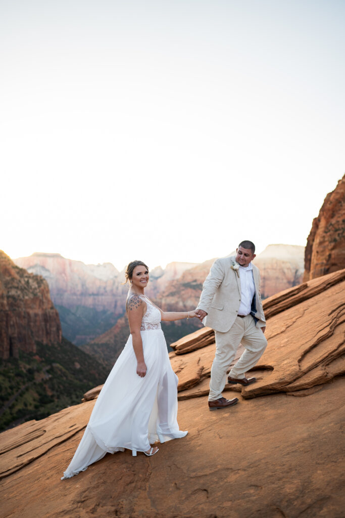 Elopement with groom leading the bride up a cliffside in Zion National Park in Utah at Canyon View Overlook at Sunset
