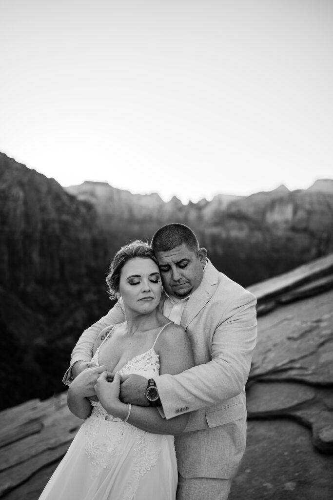 Black and white photo of elopement with Bride and Groom facing the same direction with the groom behind the bride embracing her with both arms and she has her arms wrapped around his in Zion National Park in Utah at Canyon View Overlook at Sunset