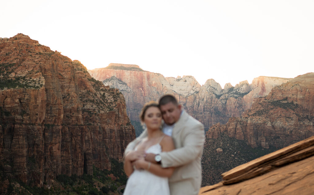 Elopement with Bride and Groom facing the same direction with the groom behind the bride embracing her with both arms and she has her arms wrapped around his in Zion National Park in Utah at Canyon View Overlook at Sunset. The couple is blurred and the background is in focus