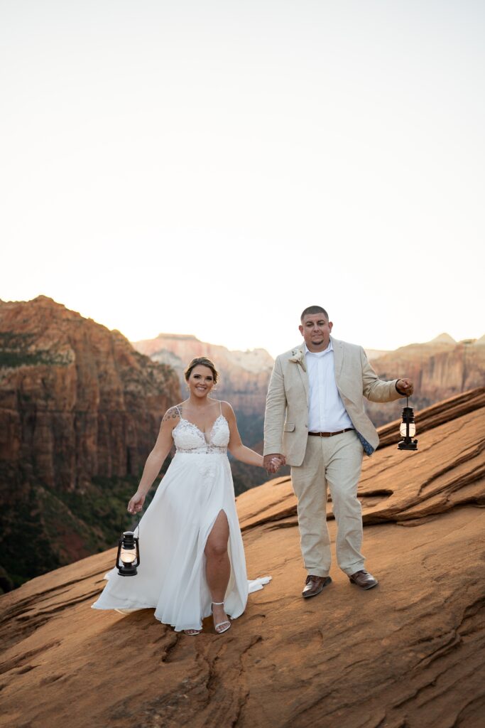 Elopement with Bride and Groom facing the the camera and holding hands in between them while holding lanterns in their other hands in Zion National Park in Utah at Canyon View Overlook at Sunset