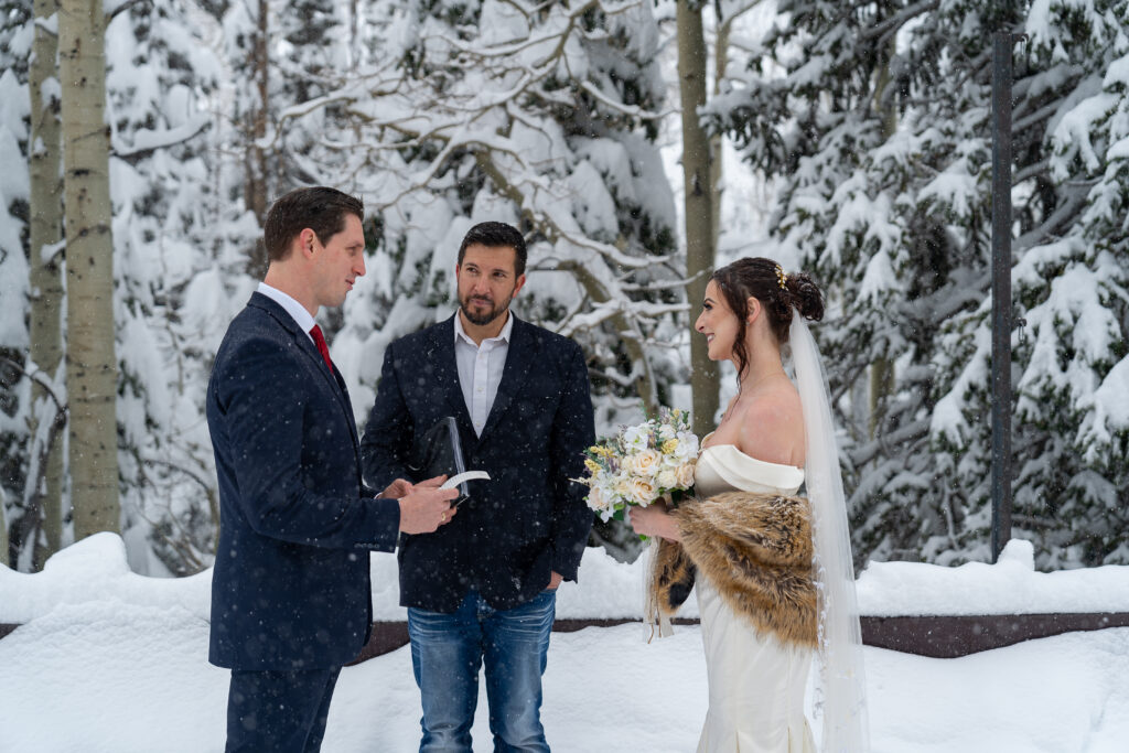 Bride and groom standing in front of their officiant during their winter elopement with snow covered pine trees in the background in Park City, Utah