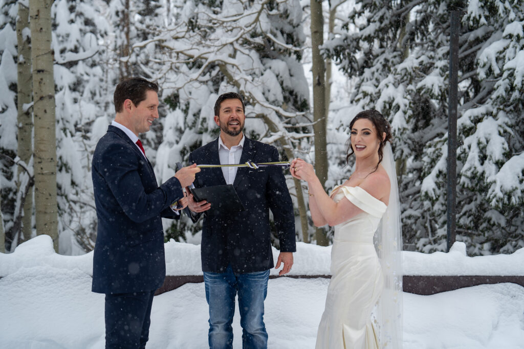 Bride and groom standing in front of their officiant during their winter elopement with snow covered pine trees in the background in Park City, Utah