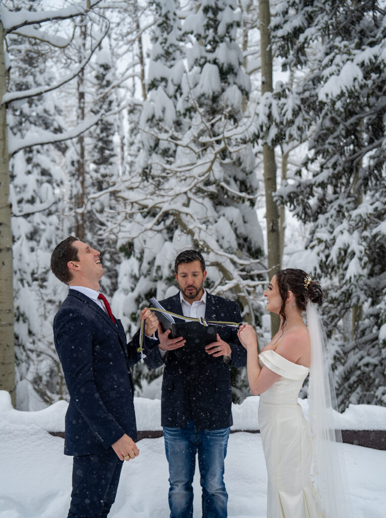 Bride and groom standing in front of their officiant during their winter elopement with snow covered pine trees in the background in Park City, Utah. Groom is laughing and bride is smiling at him.