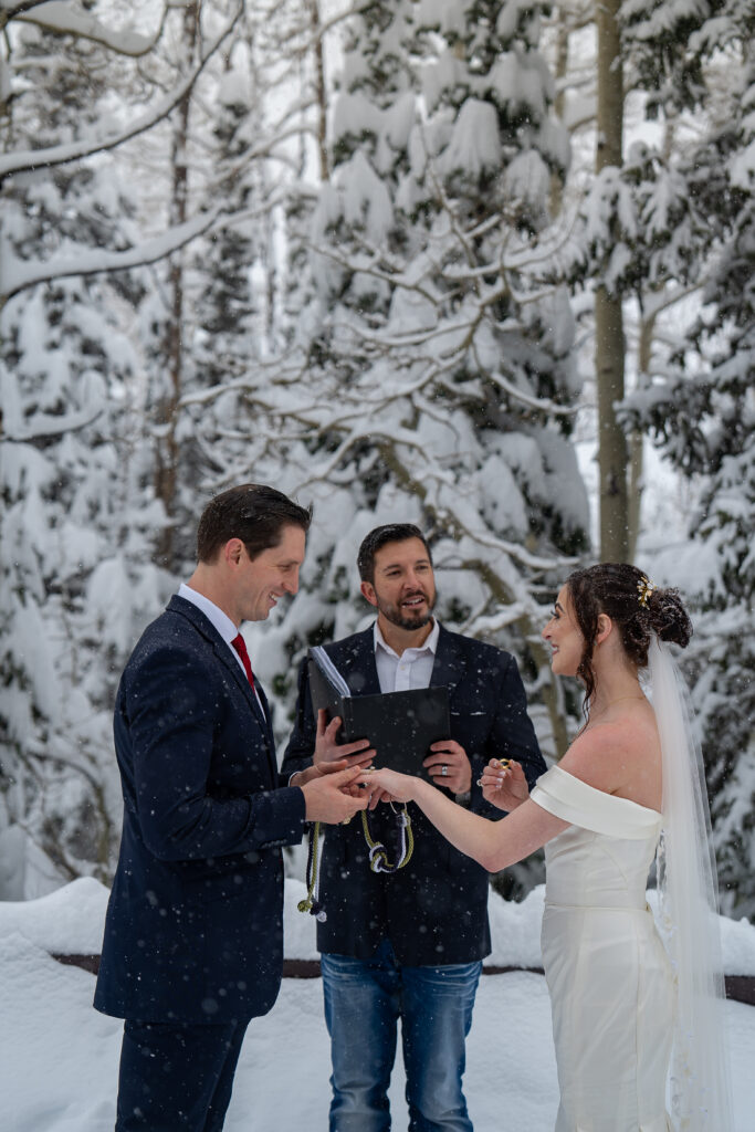 Groom putting ring on bride's finger in front of their officiant during their winter elopement with snow covered pine trees in the background in Park City, Utah