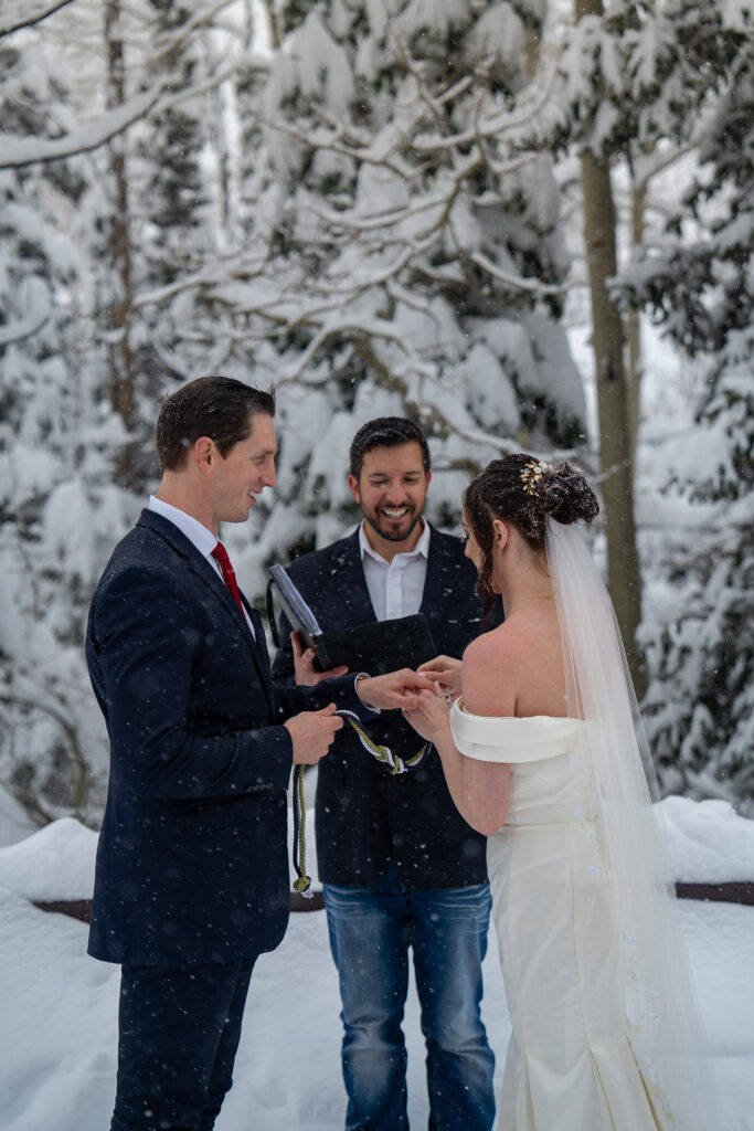 Bride putting ring on grooms finger in front of their officiant during their winter elopement with snow covered pine trees in the background in Park City, Utah