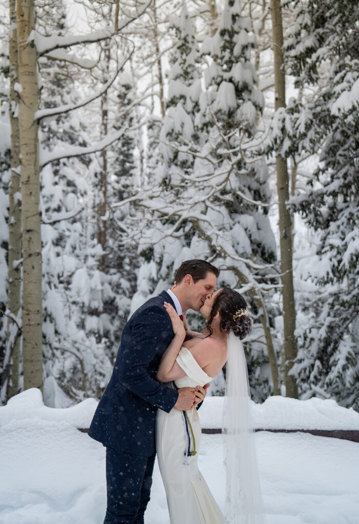 Bride and groom kissing as the snow falls on them with snow covered pine trees behind them in Park City, Utah