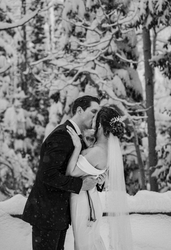 Black and white photo of bride and groom kissing as the snow falls on them with snow covered pine trees behind them in Park City, Utah
