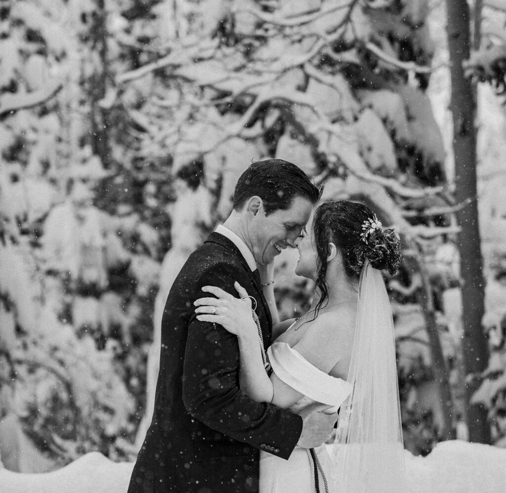 Black and white photo of bride and groom smiling while going in for a kiss as the snow falls on them with snow covered pine trees behind them in Park City, Utah