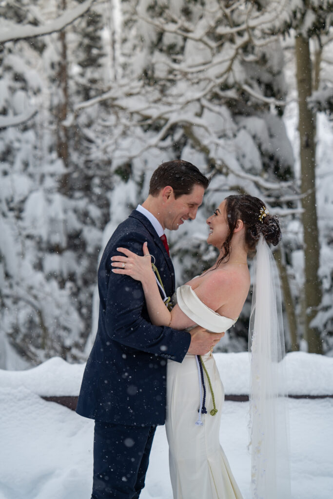 Bride and groom smiling and embracing each other as the snow falls on them with snow covered pine trees behind them in Park City, Utah