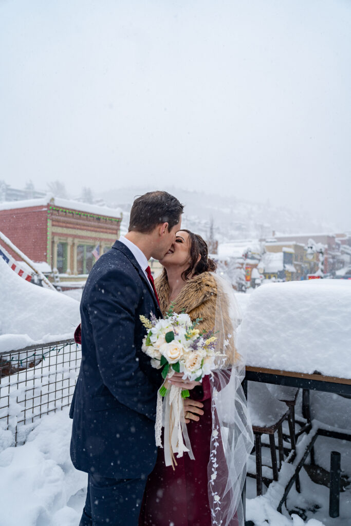 Bride and groom standing outside on the deck of the bar kissing in the snow as snow is falling in Park City, Utah