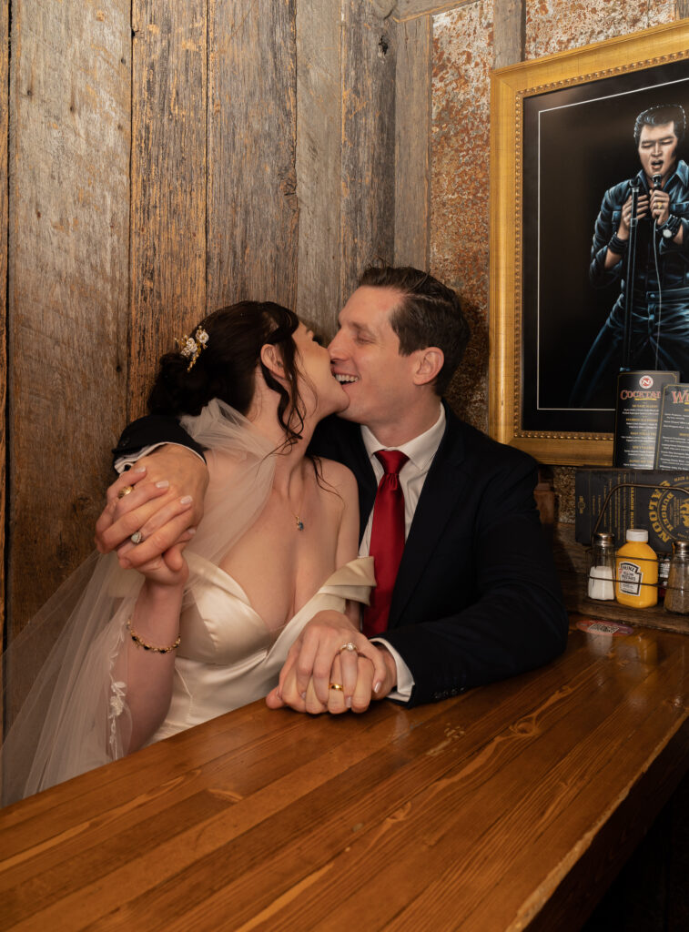 Bride and groom sitting on the same side of the table holding hands and kissing at a bar in Park City, Utah
