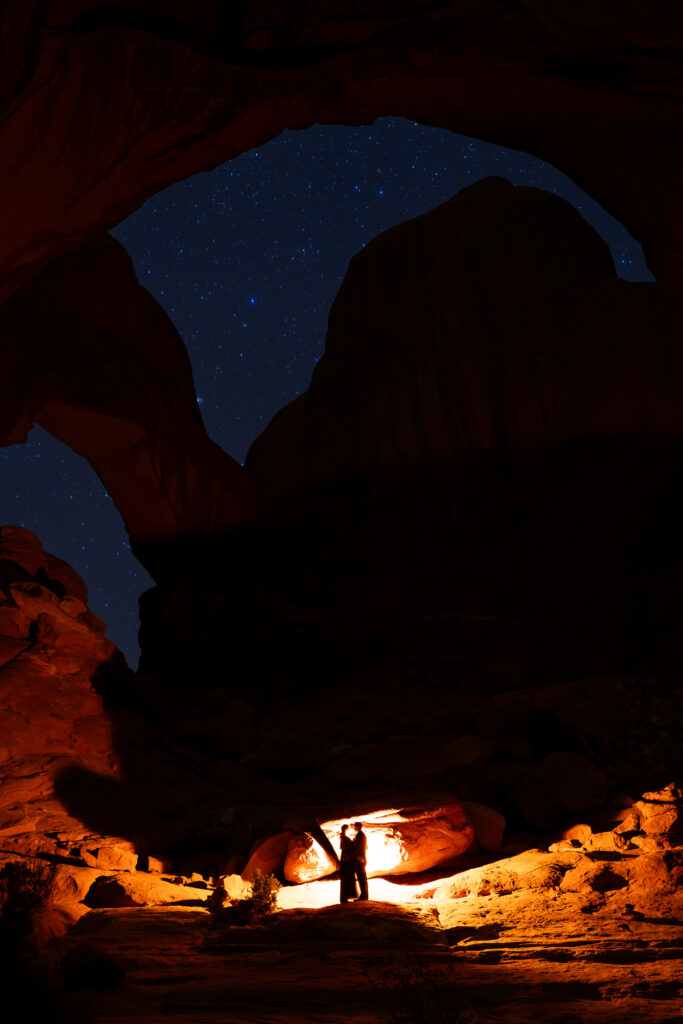 Bride and groom standing under double arch in Arches National Park at night under the stars