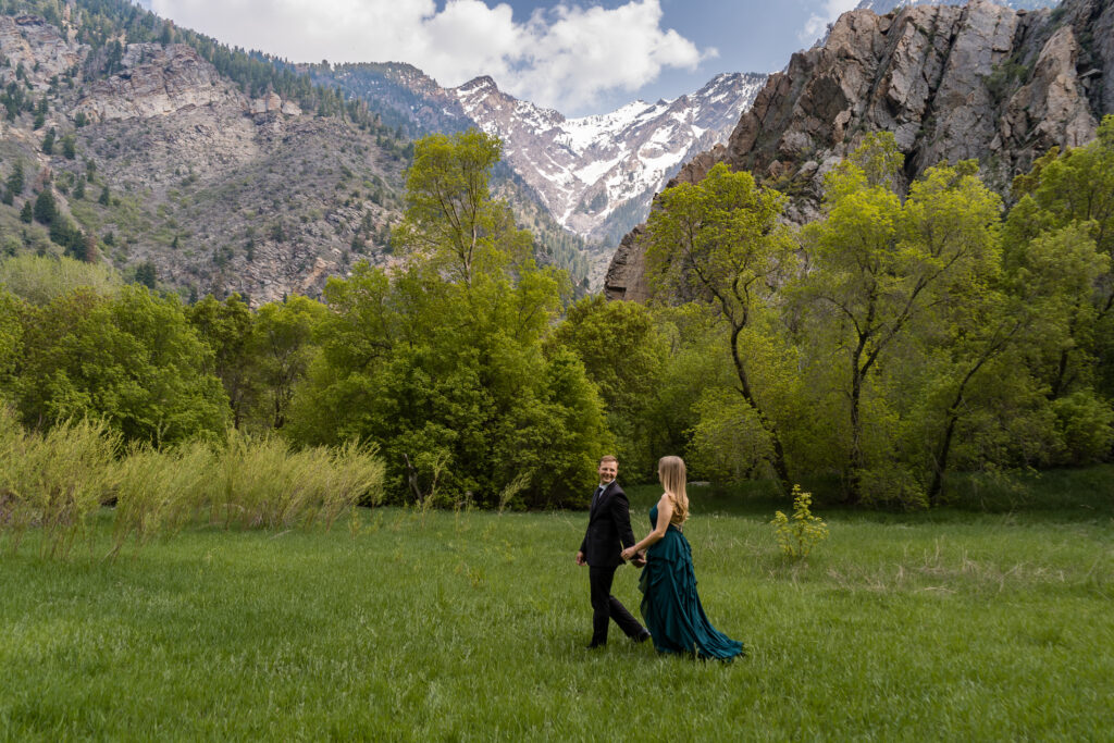 Groom leading bride across a green field in front of snow covered mountains in Salt Lake City, Utah